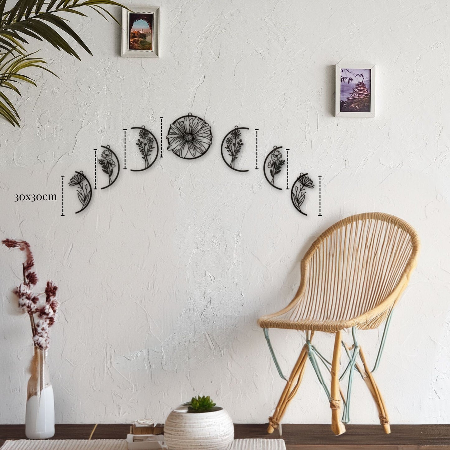 Décoration murale métal lune - ensemble de sculptures murales en métal noir de 30x30cm représentant différentes phases de la lune avec des motifs floraux, accrochées en arc sur un mur blanc texturé, à côté d'une chaise en osier et d'une plante décorative.